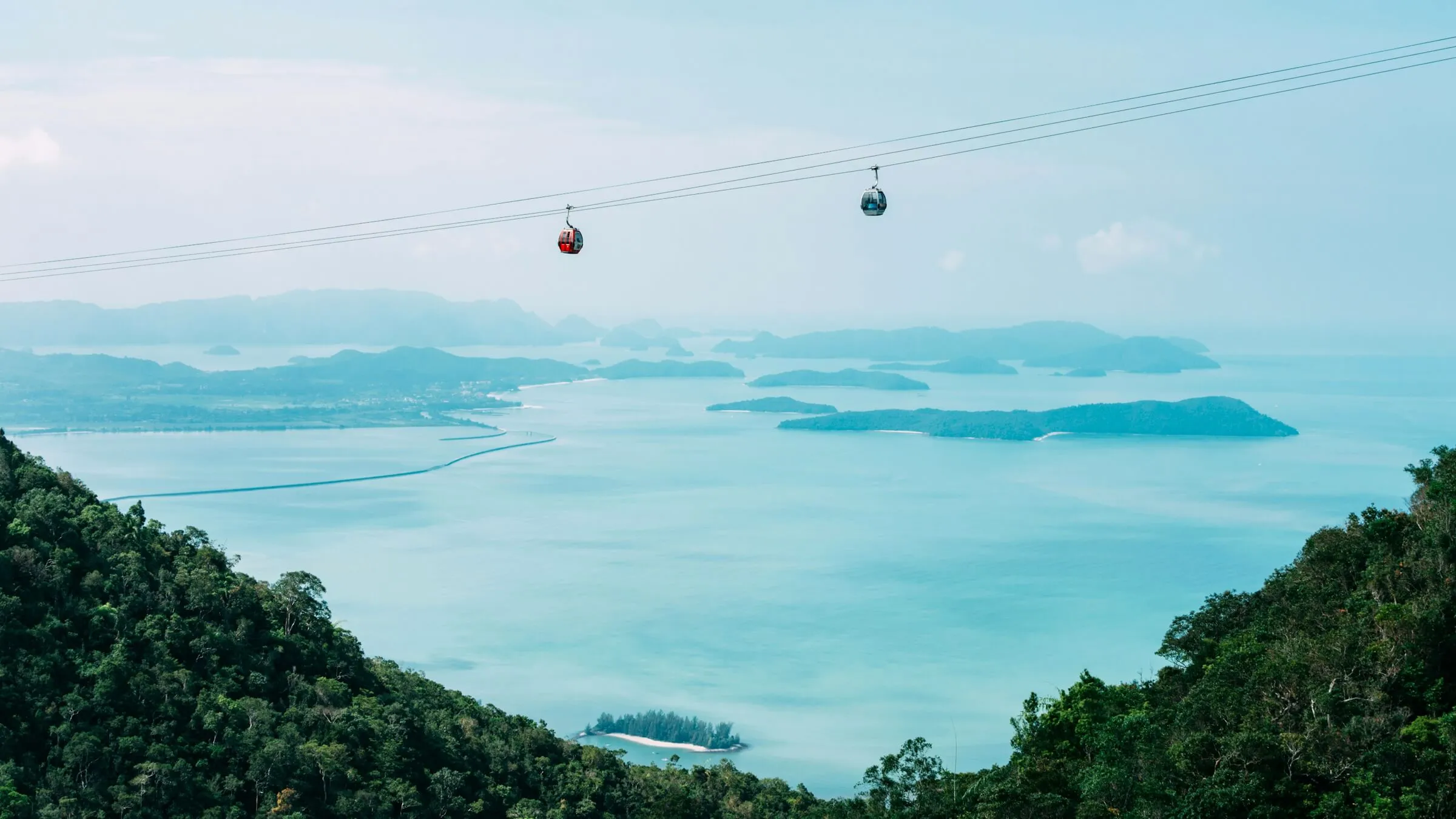 Two Langkawi Cable Car gondolas on the cable high above forested Machinchang slopes, with the turquoise Andaman Sea and scattered islands below
